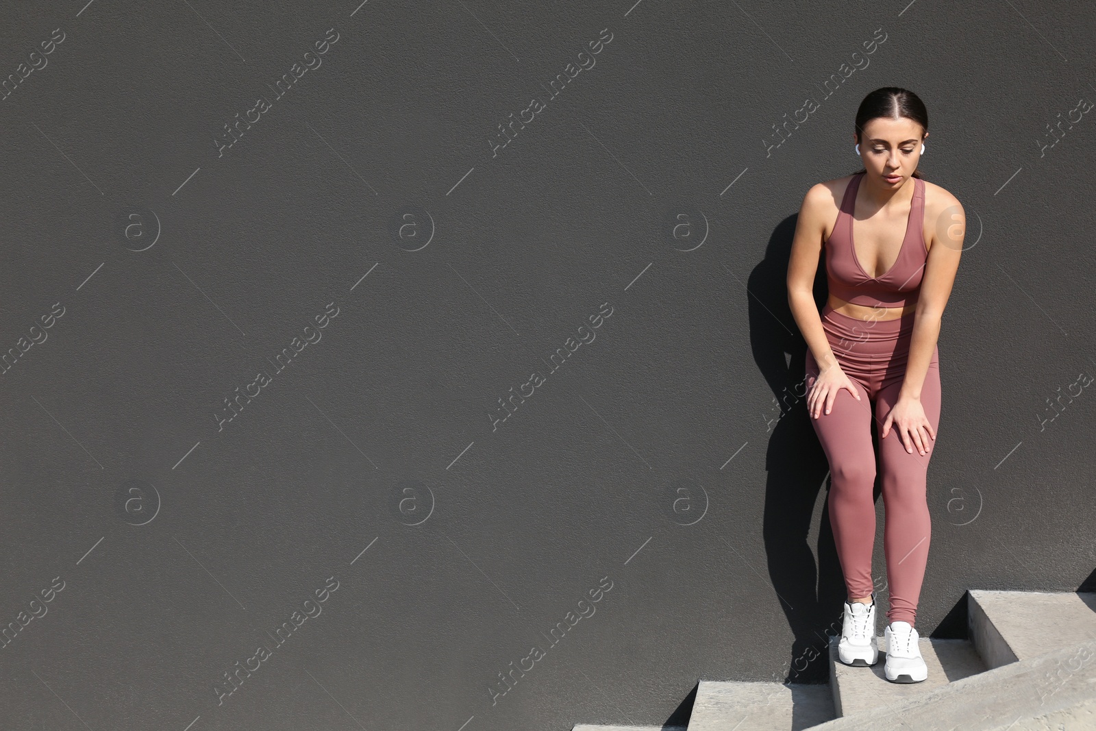 Photo of Young woman in stylish sports wear on stone stairs near grey wall. Space for text