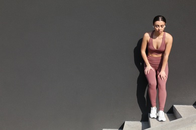 Young woman in stylish sports wear on stone stairs near grey wall. Space for text