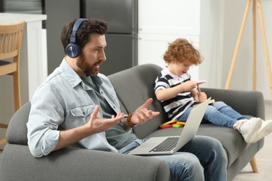 Photo of Father working remotely on laptop while his son playing with toys at home. Man having video chat with colleagues