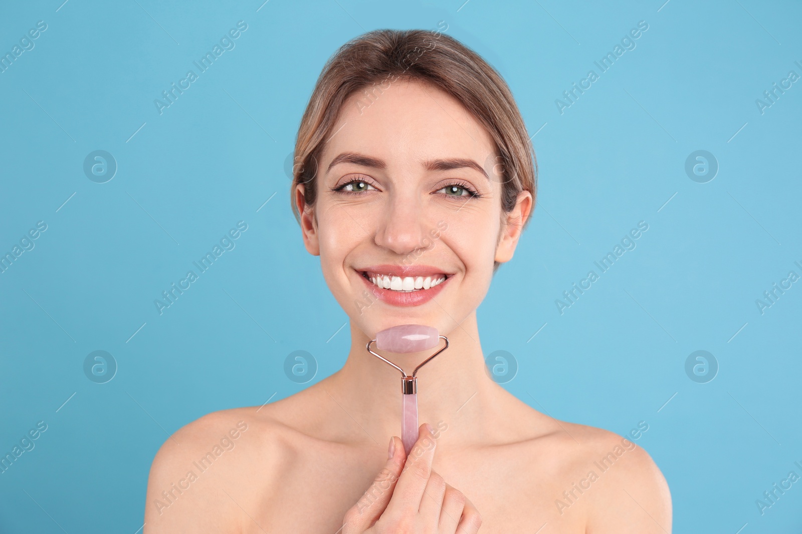 Photo of Young woman using natural rose quartz face roller on light blue background