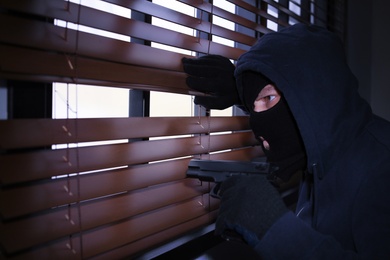 Photo of Masked man with gun spying through window blinds indoors. Criminal offence