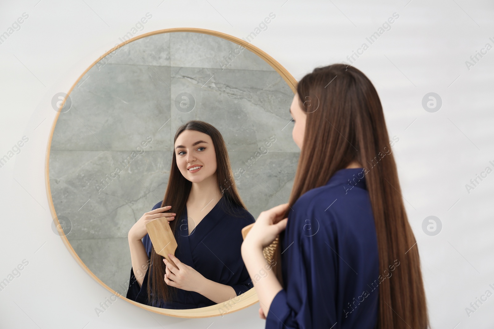 Photo of Beautiful woman in blue robe brushing hair near mirror indoors