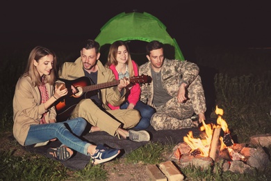Photo of Young man playing guitar for friends near bonfire at night. Camping season