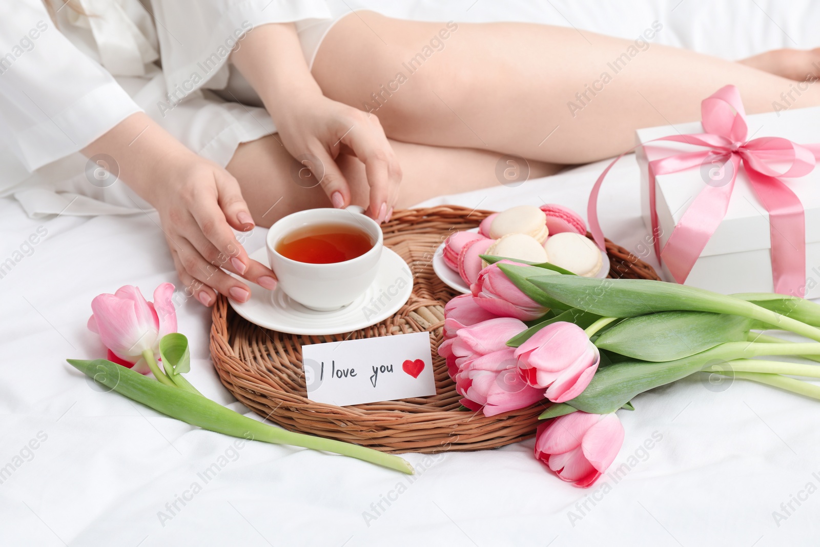 Photo of Tasty breakfast served in bed. Woman with tea, macarons, gift box, flowers and I Love You card at home, closeup