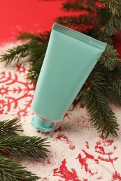 Photo of Winter skin care. Hand cream near snowflake silhouettes made with artificial snow and fir branches on red background