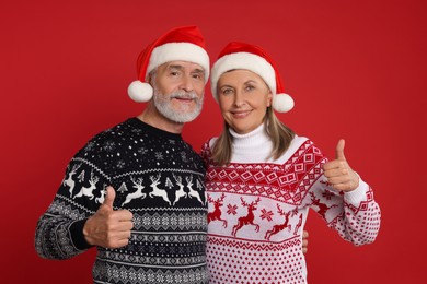 Photo of Senior couple in Christmas sweaters and Santa hats showing thumbs up on red background