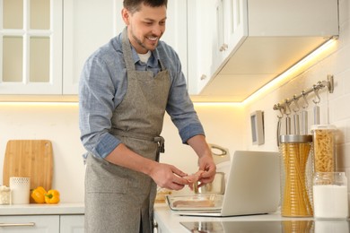 Photo of Man making dinner while watching online cooking course via laptop in kitchen