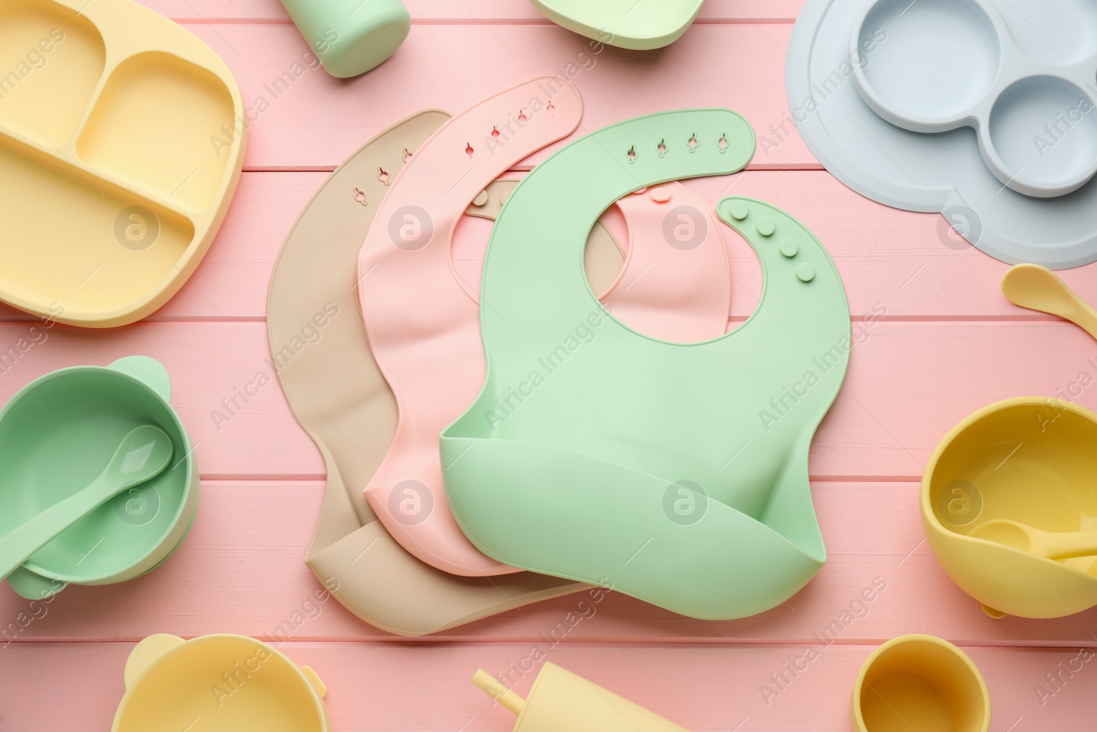 Photo of Flat lay composition with silicone baby bibs and plastic dishware on pink wooden background