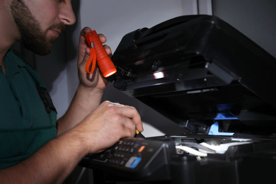 Photo of Repairman with flashlight fixing modern printer indoors, closeup