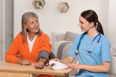 Young healthcare worker measuring senior woman's blood pressure at wooden table indoors