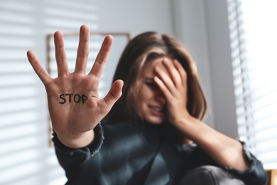 Photo of Crying young woman showing palm with word STOP indoors, focus on hand. Domestic violence concept