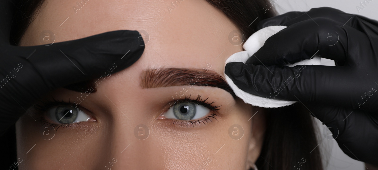 Photo of Beautician wiping tint from woman's eyebrows on light grey background, closeup