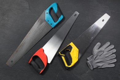 Saws with colorful handles and gloves on black textured background, flat lay