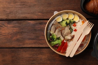 Tasty food in container with knife and fork on wooden table, flat lay. Space for text