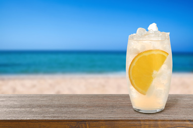 Image of Lemonade with ice cubes and orange slice on wooden table at beach, space for text