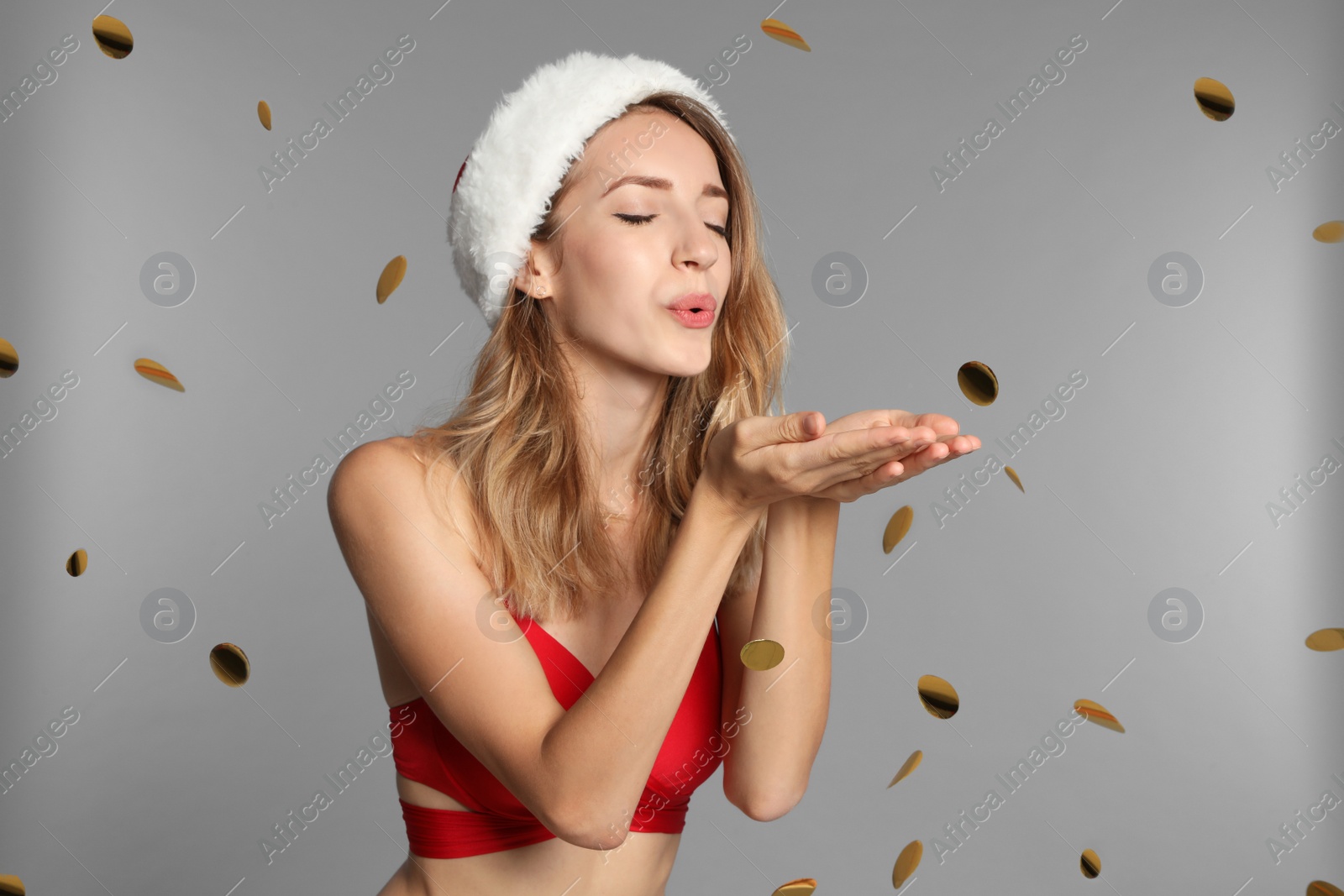 Photo of Happy young woman in red bikini and Santa hat blowing confetti on light grey background. Christmas celebration