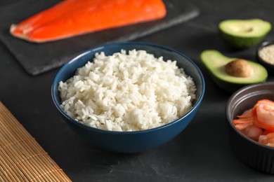 Photo of Cooked rice in bowl and other ingredients for sushi on dark table, closeup