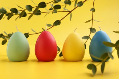 Photo of Colorful egg shaped candles and leaves on yellow background. Easter decor
