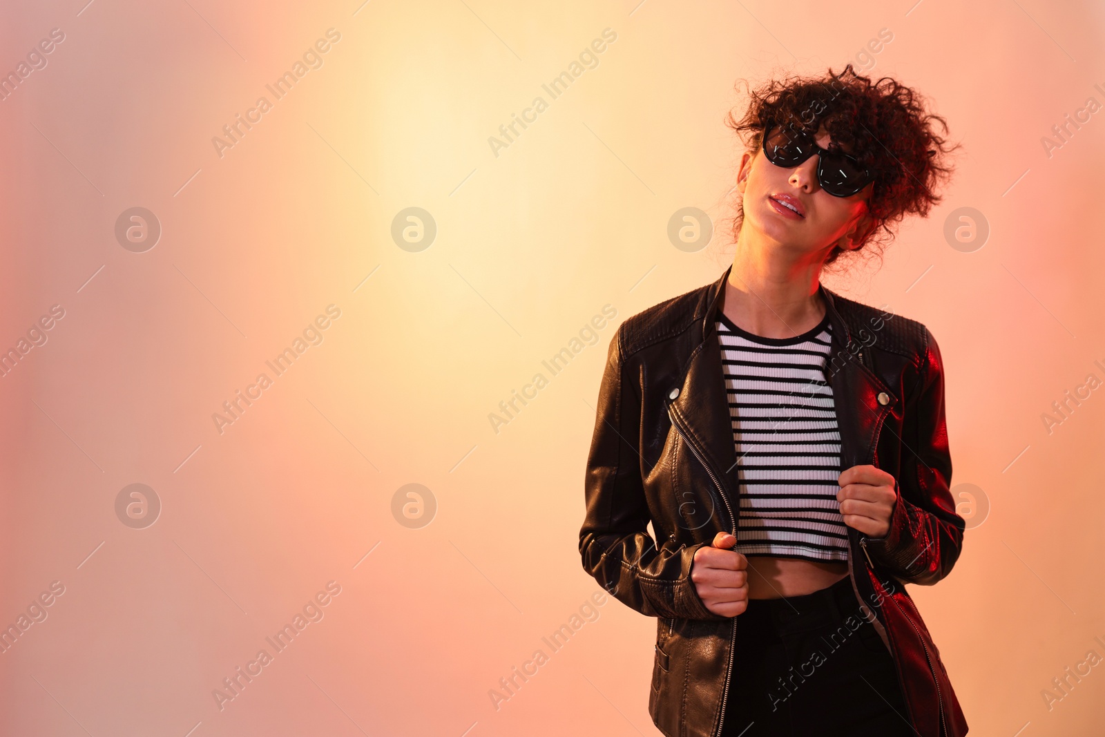 Photo of Beautiful young woman in black leather jacket and sunglasses posing on color background in neon lights. Space for text