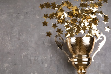 Gold trophy cup and confetti on grey stone background, flat lay. Space for text