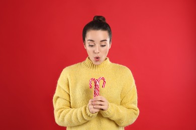 Surprised young woman in yellow sweater holding candy canes on red background. Celebrating Christmas