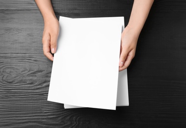 Woman holding blank paper sheets for brochure at black wooden table, top view. Mock up