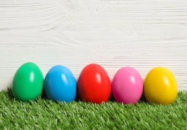 Photo of Bright Easter eggs on green grass against white wooden background