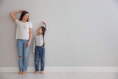 Photo of Little girl and mother measuring their height near light grey wall indoors. Space for text