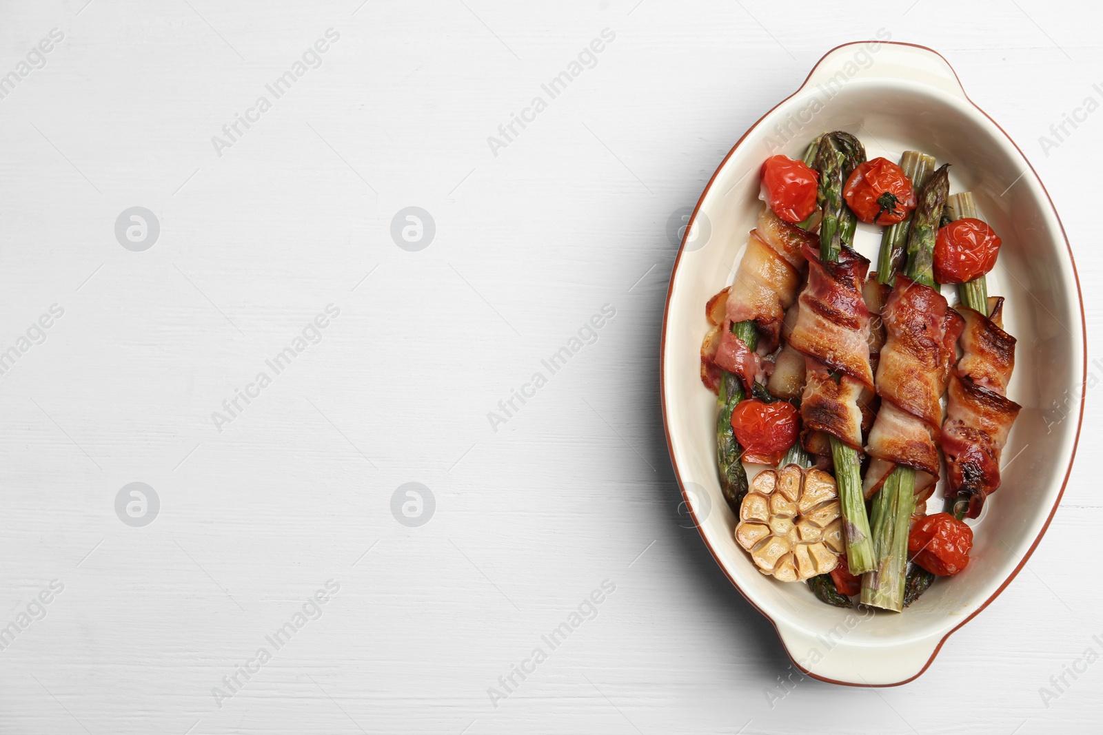 Photo of Oven baked asparagus wrapped with bacon in ceramic dish on white wooden table, top view. Space for text