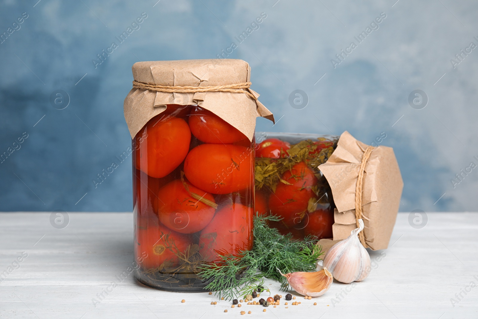 Photo of Pickled tomatoes in glass jars and products on white wooden table against blue background