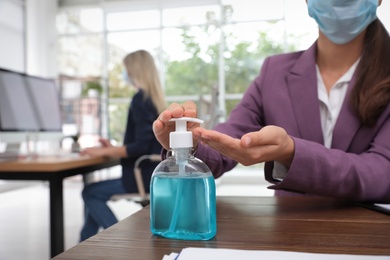 Photo of Office employee in mask applying hand sanitizer at workplace, closeup