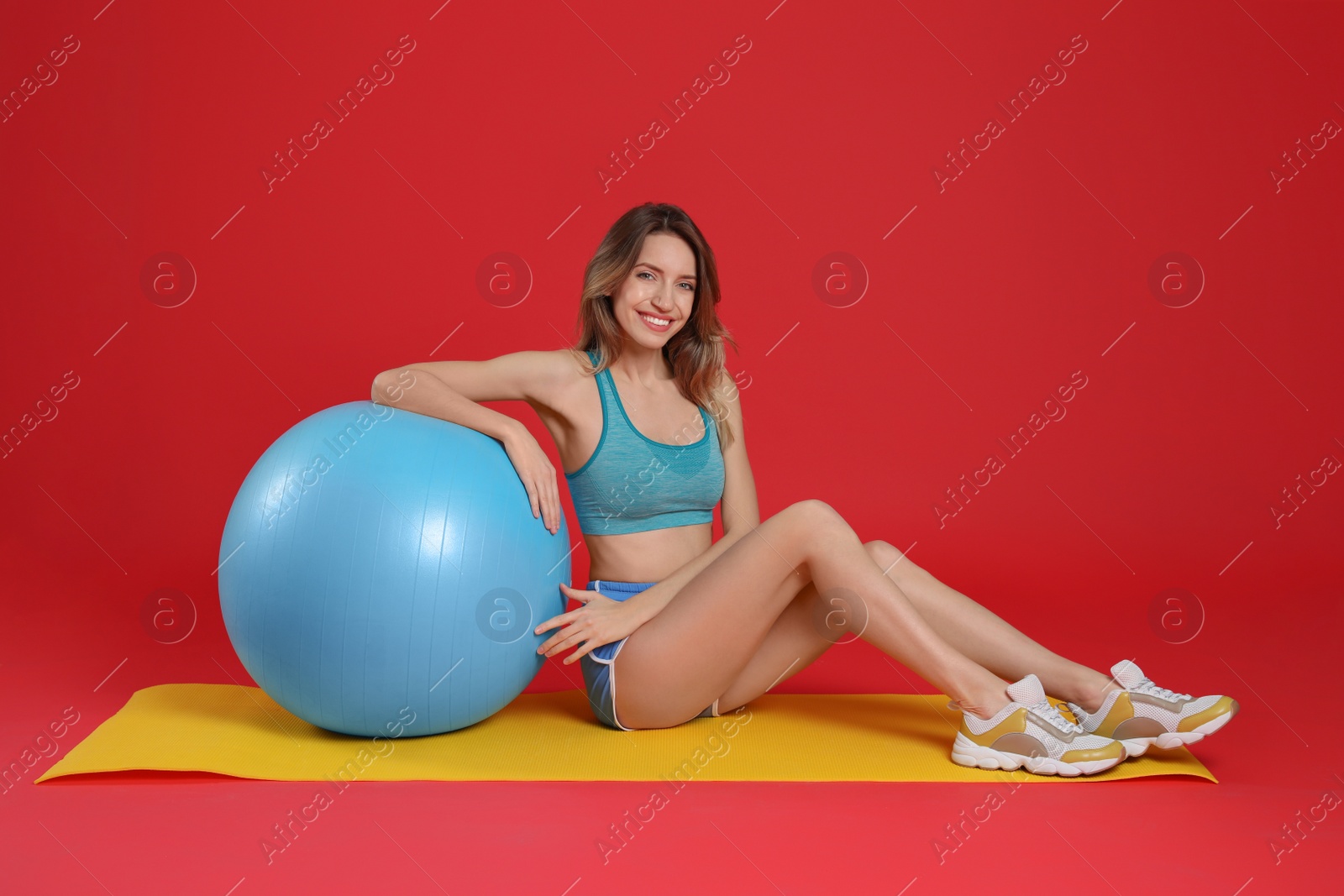 Photo of Beautiful woman sitting on yoga mat near fitness ball against red background