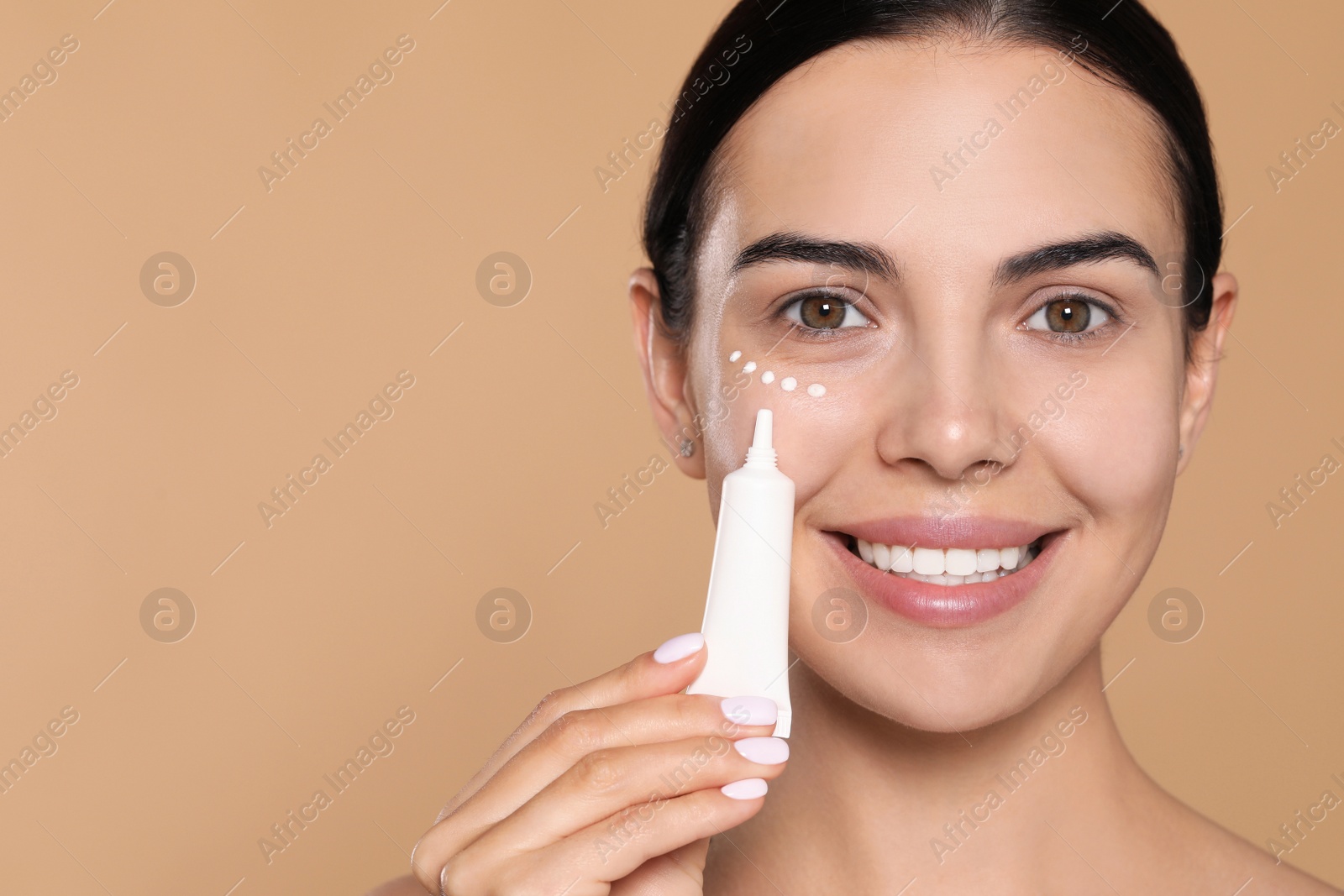 Photo of Beautiful young woman with cream on skin under eye against beige background. Space for text