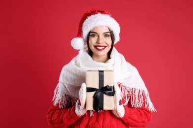 Photo of Woman in Santa hat, knitted mittens, scarf and sweater holding Christmas gift on red background