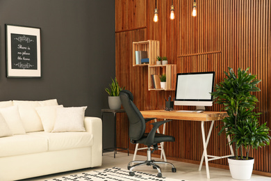 Comfortable workplace with computer near wooden wall in stylish room interior. Home office design