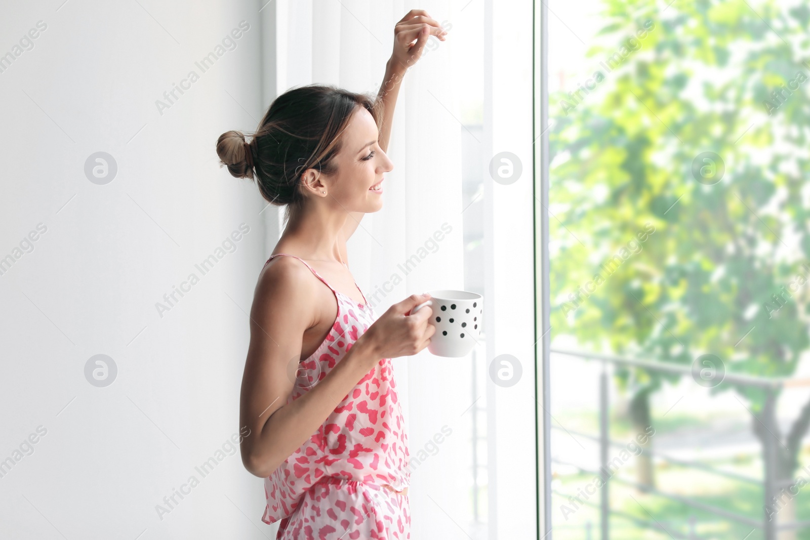Photo of Young happy woman with cup of aromatic coffee near window at home. Lazy morning