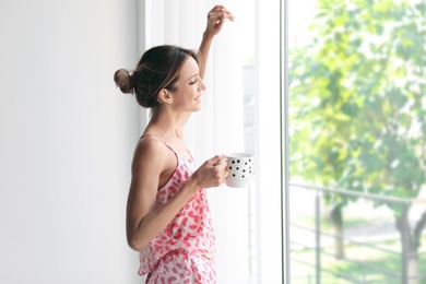 Photo of Young happy woman with cup of aromatic coffee near window at home. Lazy morning