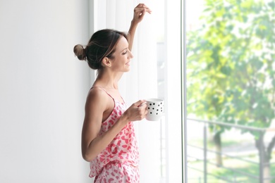 Young happy woman with cup of aromatic coffee near window at home. Lazy morning