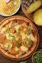 Photo of Delicious pineapple pizza and ingredients on wooden table, flat lay