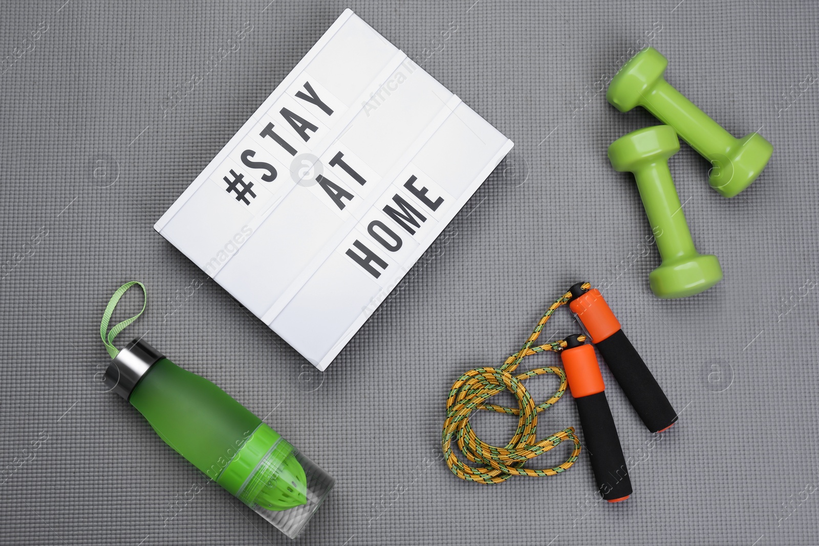 Photo of Sport equipment and lightbox with hashtag STAY AT HOME on grey yoga mat, flat lay. Message to promote self-isolation during COVID‑19 pandemic