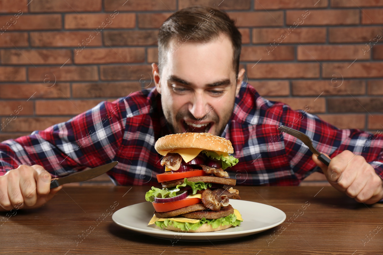 Photo of Young hungry man with cutlery eating huge burger at table