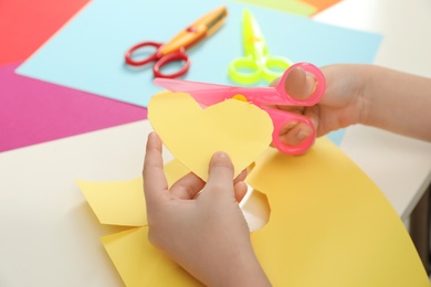 Photo of Child cutting out paper heart with plastic scissors at table, closeup