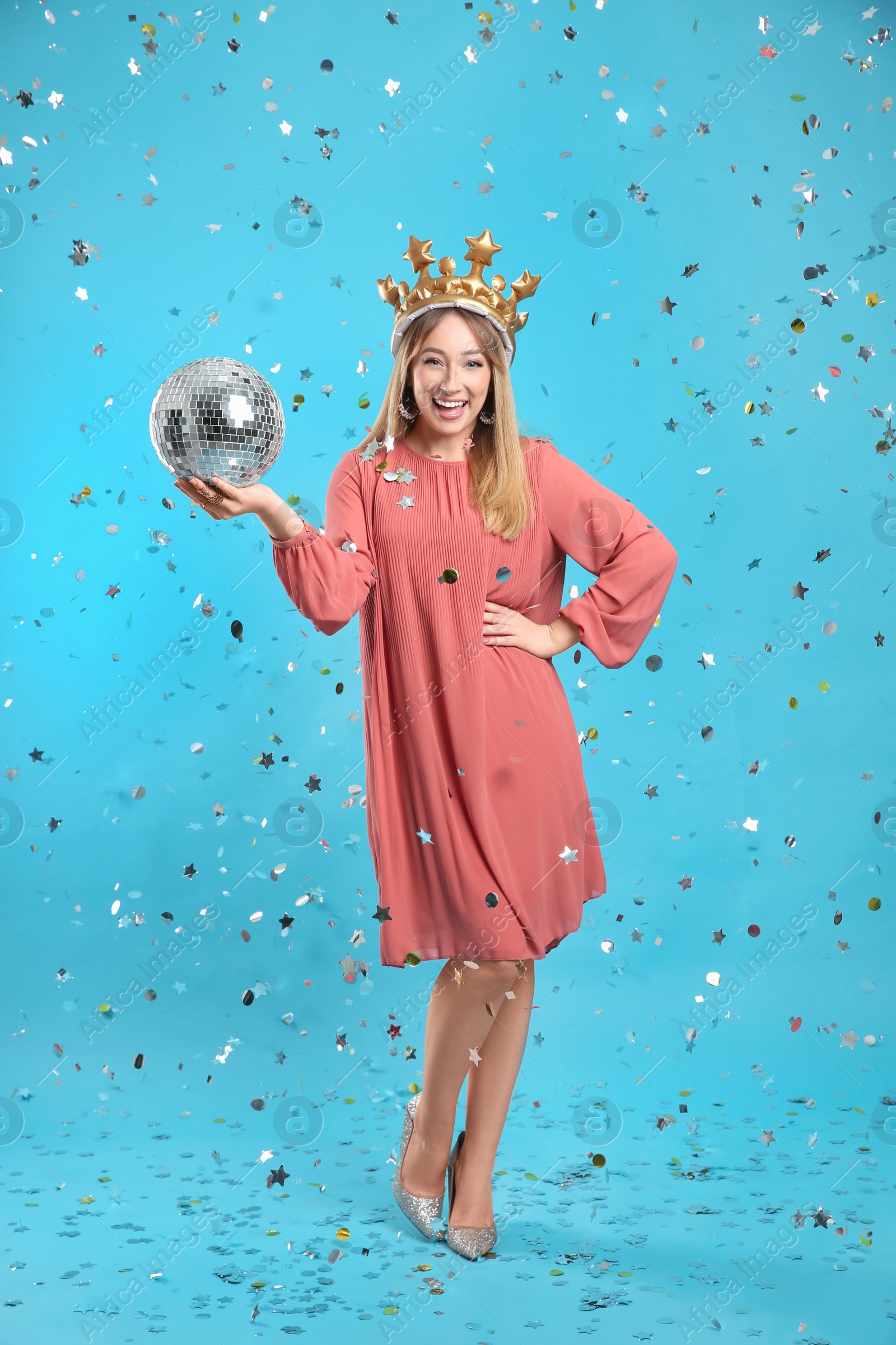 Photo of Happy young woman in party crown with disco ball and confetti on light blue background