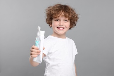 Photo of Cute little boy holding electric toothbrush and tube of toothpaste on light grey background