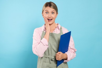 Photo of Beautiful young woman in clean apron with clipboard on light blue background