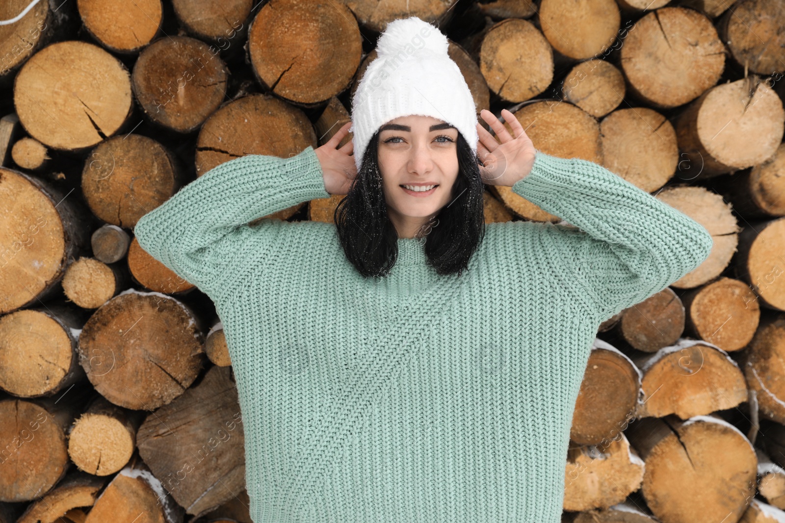 Photo of Young woman wearing warm sweater and hat near stack of firewood. Winter season