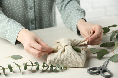 Photo of Furoshiki technique. Woman decorating gift wrapped in fabric with eucalyptus branch at wooden table, closeup