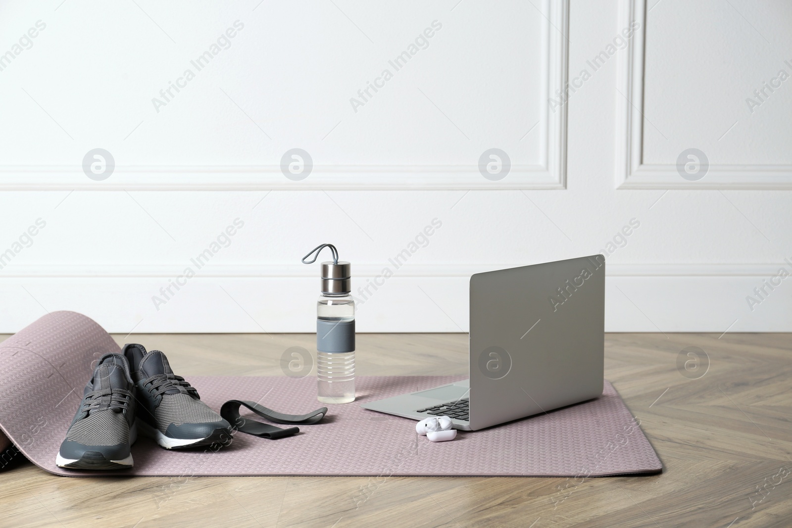Photo of Exercise mat, laptop, bottle of water, wireless earphones, fitness elastic band and shoes on wooden floor indoors