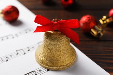 Photo of Golden shiny bell with red bow and music sheet on wooden table, closeup. Christmas decoration