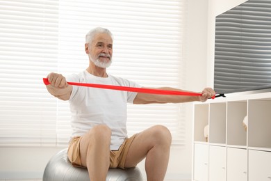 Photo of Senior man doing exercise with elastic resistance band on fitness ball at home